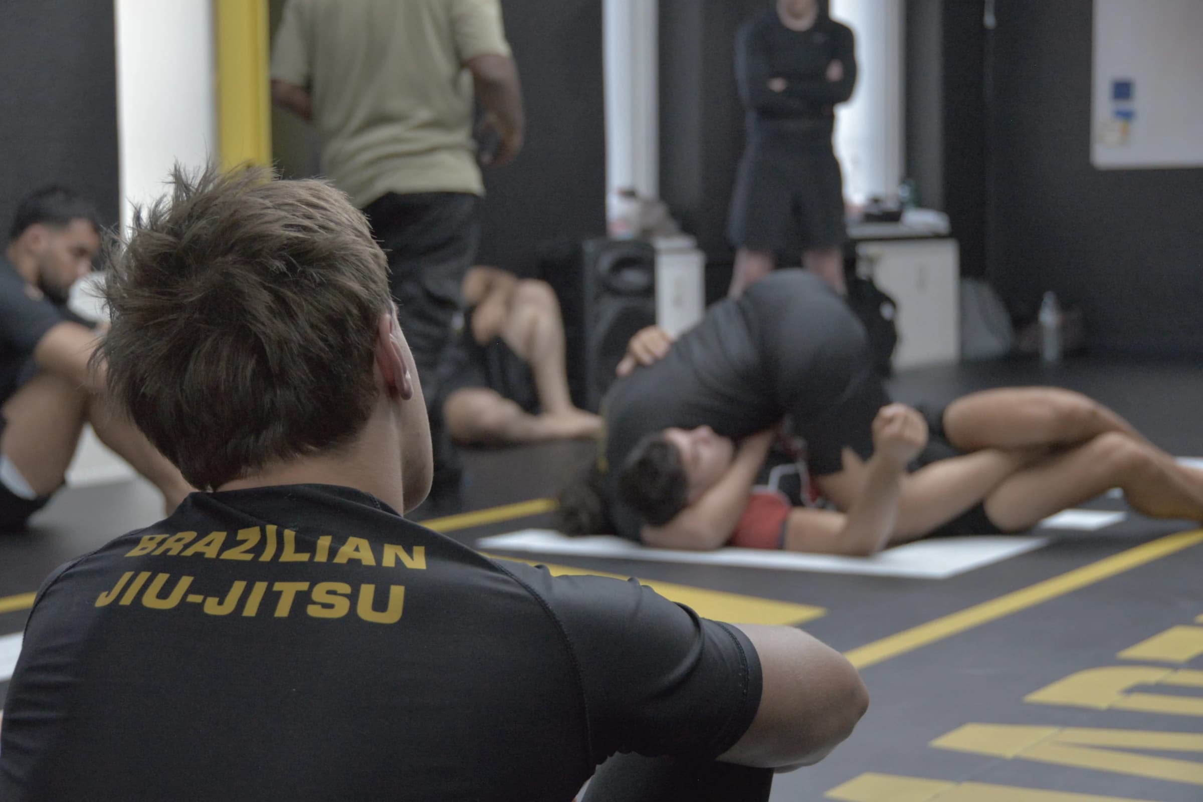 Sportler mit Brazilian-Jiu-Jitsu-Schriftzug auf dem Rücken beobachtet eine NoGi-Bodentechnik im EVLTN Gym
