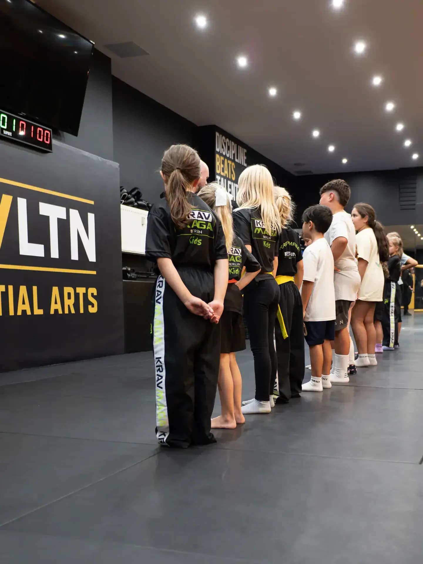A line of children in Krav Maga Kids shirts in front of the words Discipline Beats Talent at EVLTN Martial Arts in Berlin-Steglitz